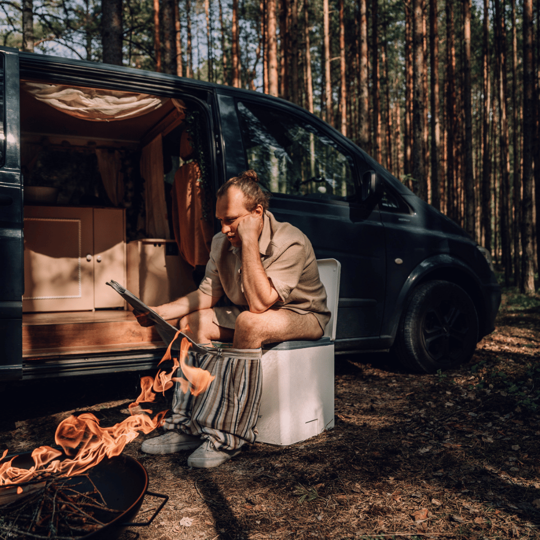 Man sitting on an Evo L composting toilet in front of his van, reading a newspaper with a fire burning nearby, in a secluded forest.