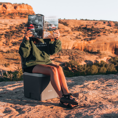 Young woman reading a newspaper while sitting on a composting toilet in a remote mountain landscape, surrounded by nature.