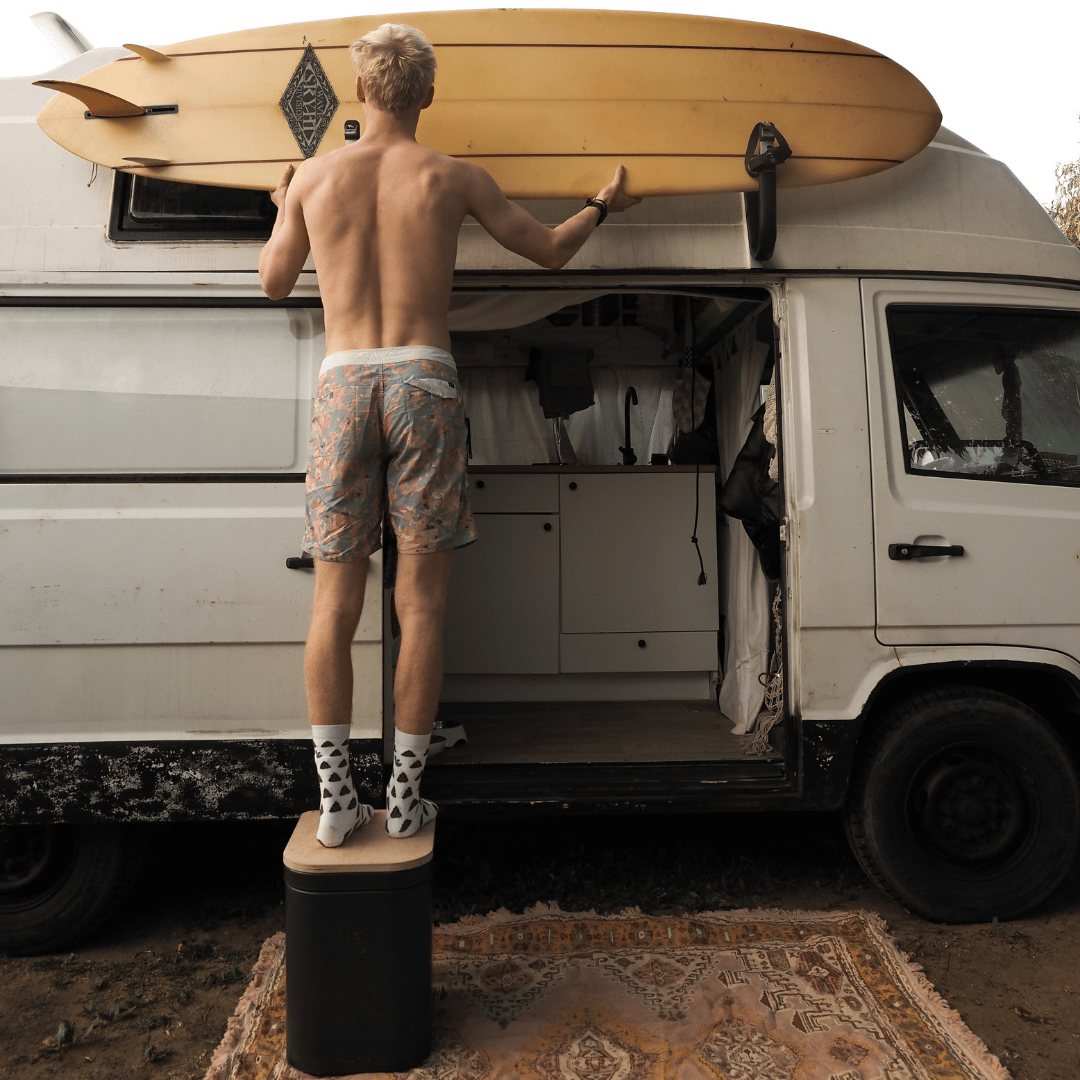 A young man stands on a Trelino Origin L composting toilet to secure a surfboard to the side of his van.