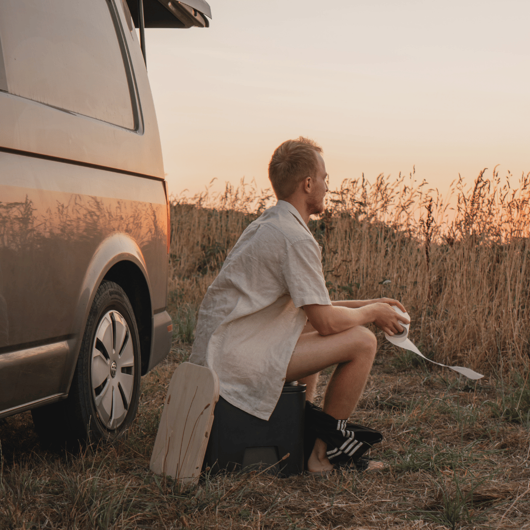 A young man sits in front of his van, watching the sunset, surrounded by an open field on a remote campsite.