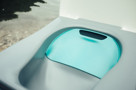 A close-up of a modern outdoor toilet seat with a light blue urine diverter resting open, set against a blurred outdoor background.