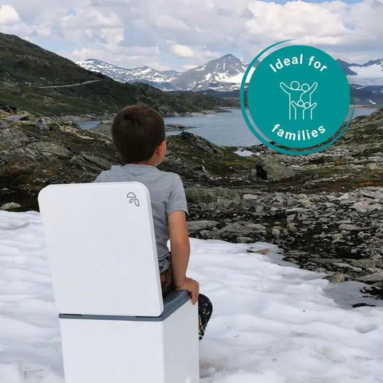 A child sits on a white outdoor portable toilet in a snowy mountain landscape near a lake, with a teal circular badge reading 'Ideal for families' in the upper right.