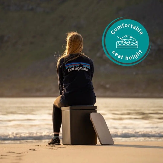A woman sits on a composting toilet on a beach, viewed from behind. 