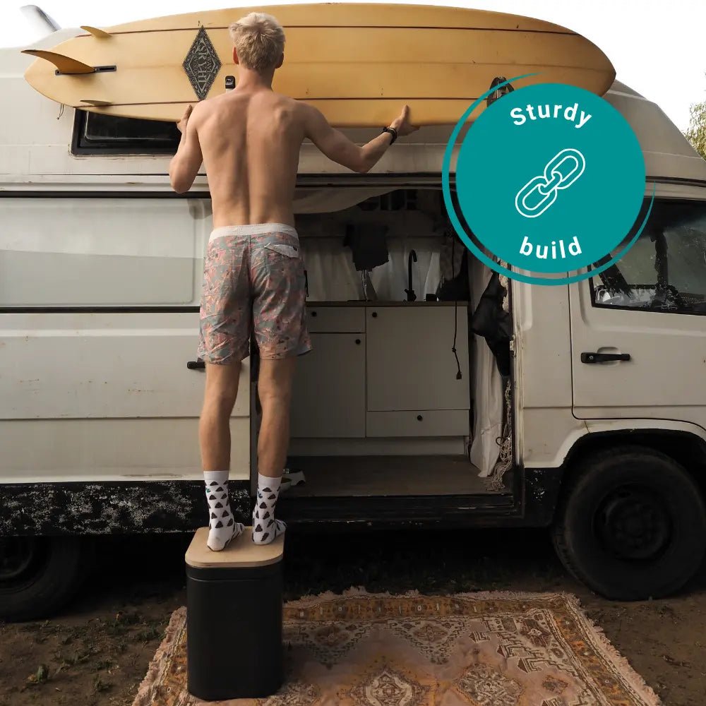 Man stands on a large Origin portable toilet in front of his camper van with a surfboard. 