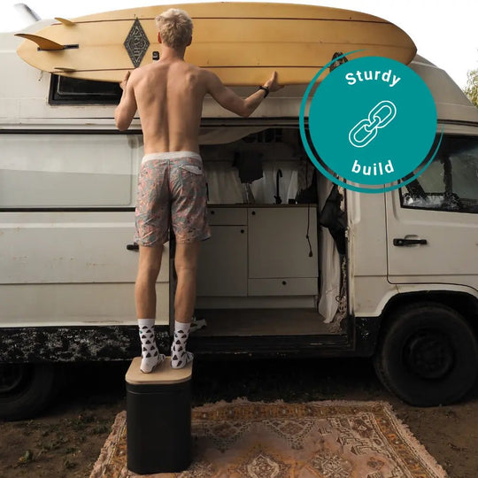 Man stands on a large Origin portable toilet in front of his camper van with a surfboard. 