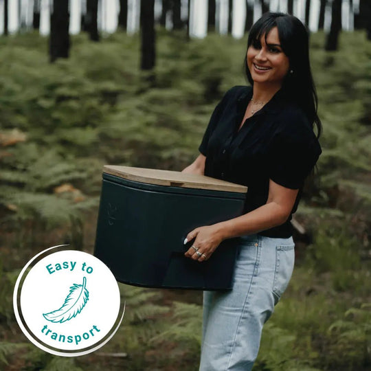 A woman stands in a forest holding an Origin M camping toilet with a light wood lid. A circular badge in the lower-left corner reads 'Easy to transport' with a feather icon.