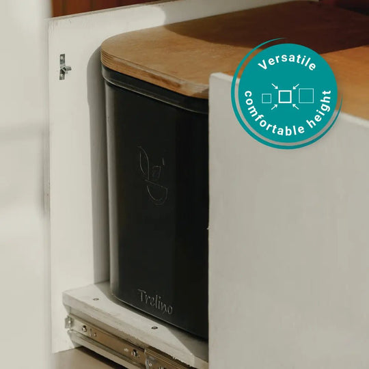 A black Trelino portable toilet with a light wood lid, shown installed inside a white cabinet. A teal circular badge on the right reads 'Versatile comfortable height'.