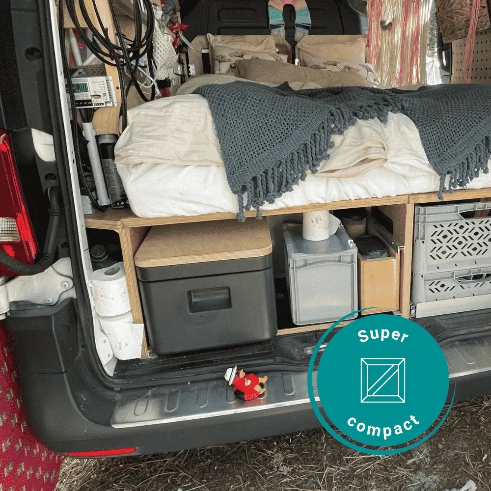 A compact camper van interior with a bed made from the rear platform, covered with white bedding and a dark gray knit throw blanket. Underneath the bed are storage bins and a Trelino portable toilet